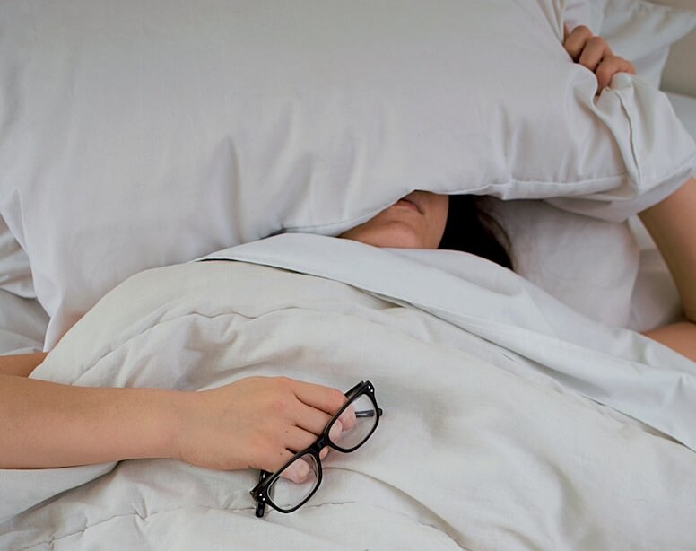 person lying on bed while covering face with pillow and holding eyeglasses